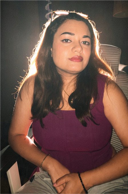 A woman with long dark hair, wearing a purple tank top and sunglasses on her head, poses for a photo with warm lighting behind her.