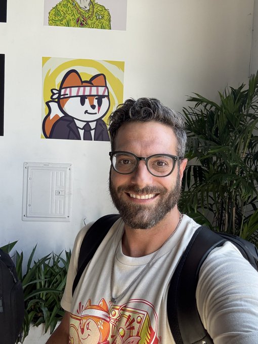 A smiling bearded man with glasses wearing a graphic t-shirt takes a selfie indoors, with anime-style artwork pinned to the white wall behind him.