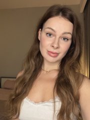 A young woman with long wavy brown hair and a white top poses for a portrait photo, wearing a delicate necklace with natural makeup.