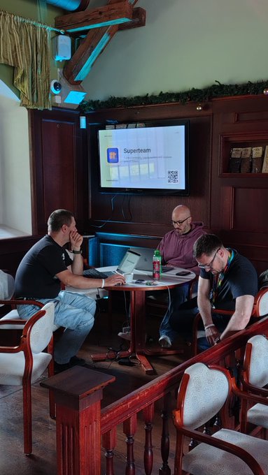 Three men gather around a table in a wood-paneled pub-style room, engaged in a focused discussion with open laptops and drinks, while a projector screen displays a "Superteam" presentation featuring a QR code—capturing a productive "build station" working session in action.