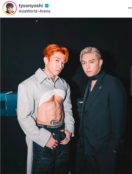 An Instagram post from user 'tysonyoshi' tagged at 'AsiaWorld-Arena' shows two stylishly dressed men posing together, one with orange hair wearing a cropped shirt revealing his abs, and the other with blonde hair in a black suit.