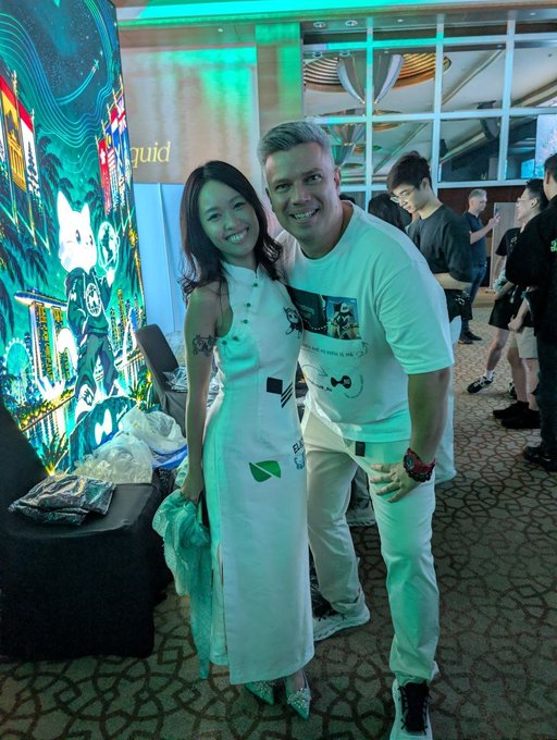 A smiling woman in a white floral-patterned dress poses alongside a man wearing a white graphic t-shirt and light pants, standing near a colorful illustrated display at what appears to be an event venue with the partial text "luid" visible in the background. Their enthusiastic poses and the festive setting suggest they're ready to embrace another memorable experience together.