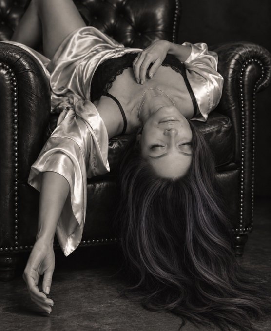 A black and white photo of a woman wearing a silk robe and lace lingerie lying upside down on a dark leather tufted Chesterfield chair.