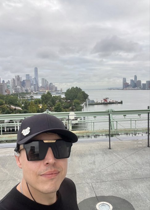 A selfie of a man wearing a black cap and large square sunglasses, with the New York City skyline and Hudson River visible in the background.