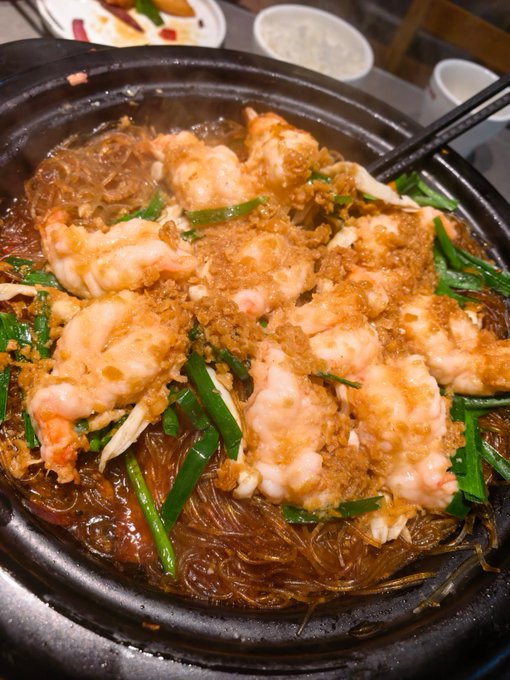 A hot pot dish featuring shrimp, glass noodles, green onions, and garlic in a savory sauce, served in a black clay pot.