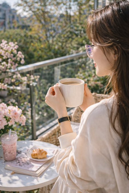 A woman with long dark hair, wearing glasses and a white blouse, holds a coffee mug at an outdoor café table with a pink drink and pastry, with blooming flowers in the background.