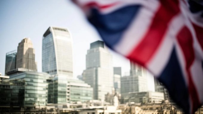 A blurred Union Jack flag waves in the foreground with the London skyline, including skyscrapers, visible in the background.