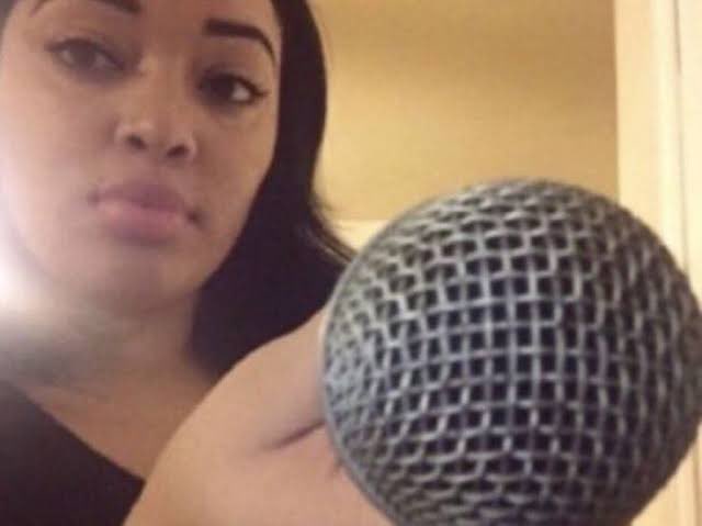 The image features a woman holding a microphone with her arm outstretched. The background of the photo shows a wall that appears to be white or tan in color. There are no other objects or people visible in the image, and the focus is solely on the woman and the microphone she's holding.