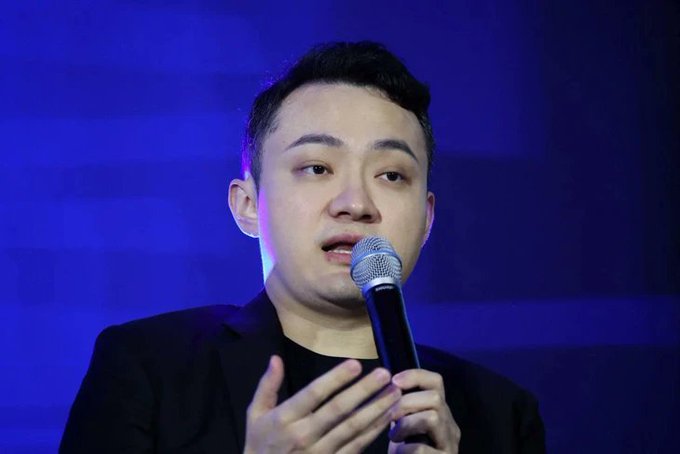 A man with short dark hair in a black blazer speaking into a microphone against a blue background, gesturing with his hand.