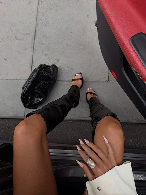 A first-person perspective photo of a person stepping out of a red car, wearing black knee-high gladiator sandals, with long manicured nails and a large ring visible on their hand, and a black bag placed on the sidewalk.