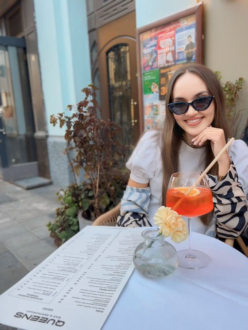 A smiling young woman in sunglasses and a white patterned top sits at an outdoor café table with an orange Aperol Spritz cocktail and a menu, with a street and poster-covered wall in the background.