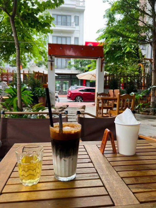 Một ly cà phê sữa đá trên bàn gỗ tại quán cà phê ngoài trời, bên cạnh một cốc nước và một hộp khăn giấy, khung cảnh cây xanh và đường phố phía sau. Bài đăng gợi liên tưởng đến việc nhâm nhi cà phê trong lúc lướt X và kết nối với những người “đồng gu,” dù “coin đỏ” vẫn vui vẻ chấp nhận “Ổn thôi.”