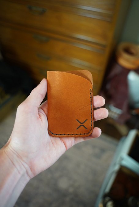 A hand holds a small, tan leather wallet with a minimalist 'X' logo on the front, set against a blurred background of a wooden dresser.