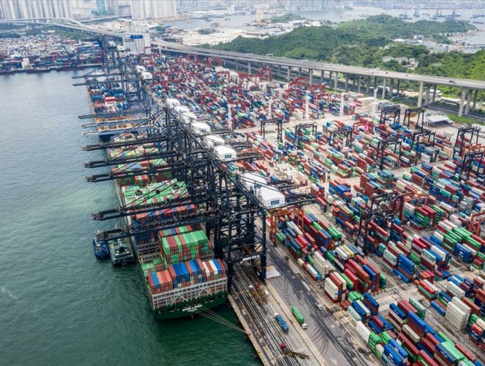 Aerial view of a busy container shipping port with cranes loading cargo ships, with a city skyline and highway in the background.