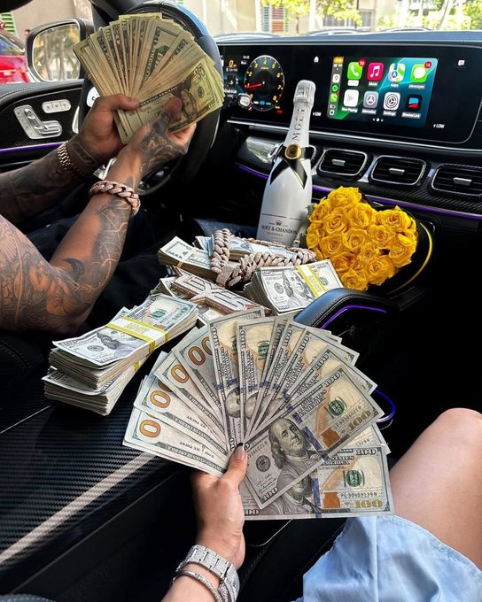 Inside a luxury car's cockpit, tattooed arms fan out stacks of hundred-dollar bills while a bottle of Moët & Chandon champagne and a bouquet of yellow roses sit beside the center console, with the dashboard screen displaying Apple CarPlay icons—visually reinforcing the post's boast to "test your luck" by flaunting lavish crypto gambling spoils.