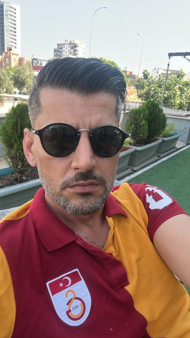 A man wearing round sunglasses and a maroon and yellow Galatasaray soccer jersey (featuring a crescent moon and star patch with "3") poses for a selfie on a rooftop terrace. The post references his fitness check continuing until reaching weights like "@btcyle," aligning with the sporty, confident vibe of his Galatasaray team attire.