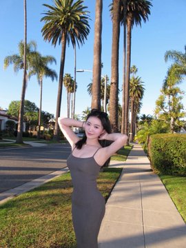 A young woman in a fitted gray dress poses on a palm tree-lined sidewalk in sunny California, hands behind her head, looking confidently at the camera—her warm, inviting demeanor playing into the playful, flirtatious caption about whether her followers would kiss her if she asked.