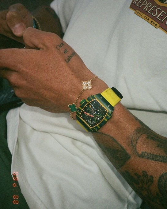 A close-up of a person's wrist showcasing a striking yellow and black watch alongside a delicate flower charm bracelet, with hand tattoos reading "Ora et Labora" and "REPRESENT" visible on the shirt. The bold timepiece and detailed accessories give off a confident, stylish vibe fitting the "we go again gm 𝕏" morning motivation.