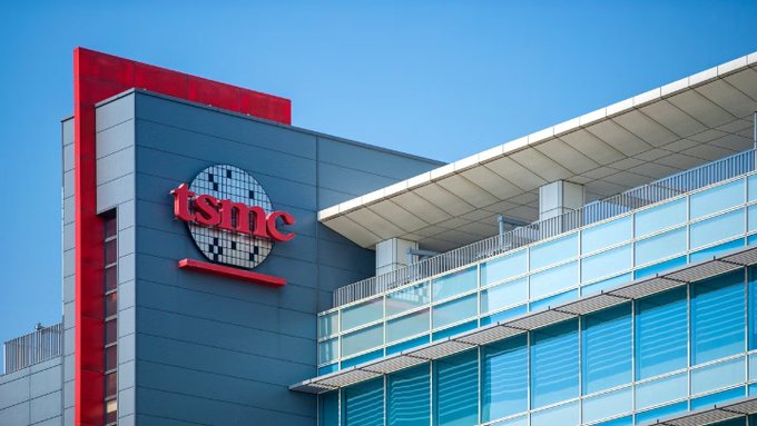 An exterior view of a TSMC (Taiwan Semiconductor Manufacturing Company) building featuring a circular emblem mounted on a dark gray facade, with red architectural accents and modern glass windows visible under a clear blue sky.