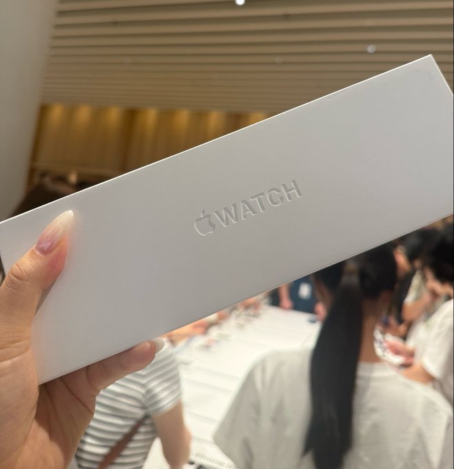 A hand holding a white Apple Watch box in what appears to be an Apple Store, with customers in the background.