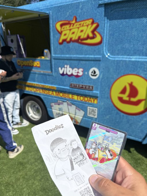 A hand holds a smartphone displaying a Doodles NFT and a printed card, with a denim 'Collector Park' branded truck visible in the background.