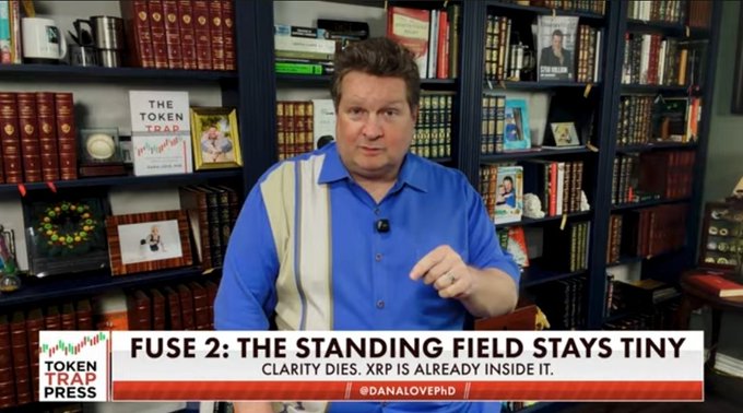 A man in a blue polo shirt stands in front of bookshelves displaying various books and memorabilia. The lower banner reads "FUSE 2: THE STANDING FIELD STAYS TINY" with a subtitle "CLARITY DIES. XRP IS ALREADY INSIDE IT," alongside the "Token Trap Press" logo and the handle "@DANALOVEHD."