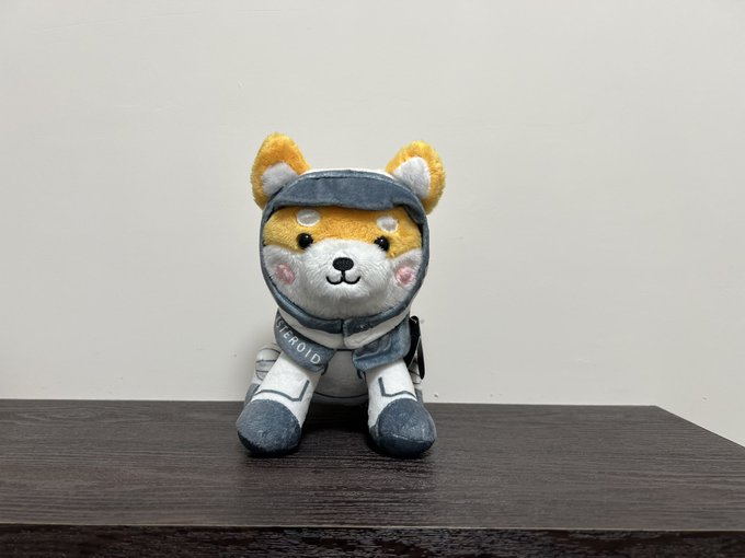 A plush Shiba Inu dog wearing a gray astronaut-style spacesuit with "EEROID" visible on the chest, sitting on a dark wooden surface. The cute "Asteroid Shiba" toy matches the post's affectionate description of an adorable space-themed Shiba character.