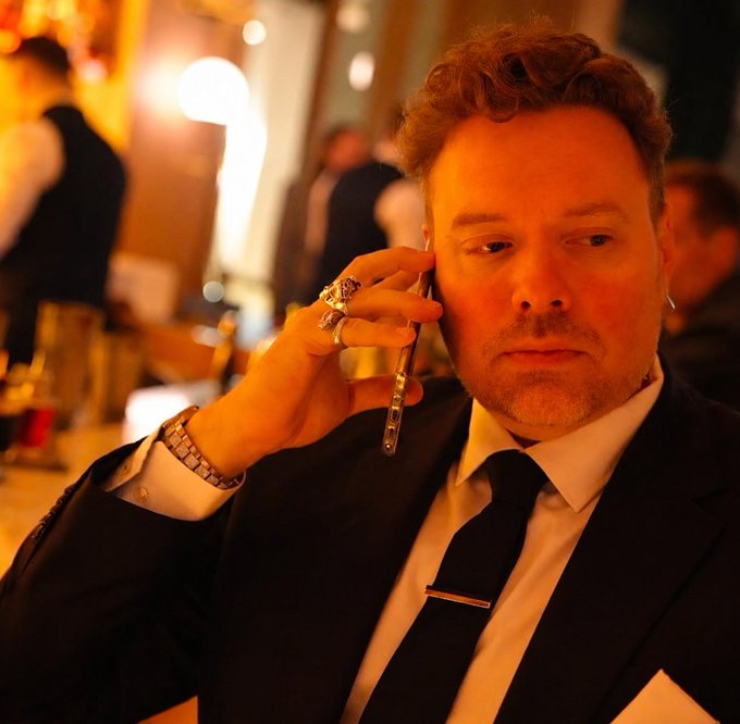 A red-haired man in a dark suit and tie holds a phone to his ear while wearing multiple rings and a wristwatch, photographed with warm, dramatic lighting at what appears to be a formal event.