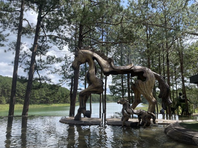 A large wooden sculpture of a horse and foal standing in shallow water, set against a backdrop of tall pine trees and a lake.