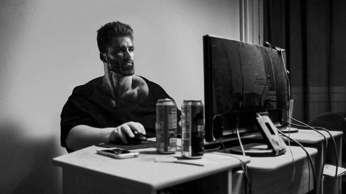 A black-and-white photograph of a bearded man with face tattoos working intently at a computer setup, with two beverage cans on the desk beside the monitor.