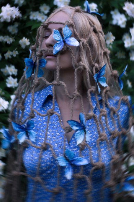 A woman with long blonde hair styled with a netting of ropes and adorned with blue butterflies poses with closed eyes against a background of white flowers, wearing a blue floral dress.
