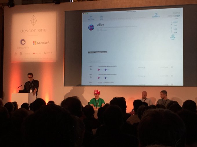 A presenter stands at a podium during the DevCon One conference (sponsored by Microsoft and ConsenSys), while a large screen displays the Mist wallet interface showing an account named "Alice" with a balance of approximately 1,851 ETH and recent transactions, as panel guests including Ethereum founders sit on stage before an audience.