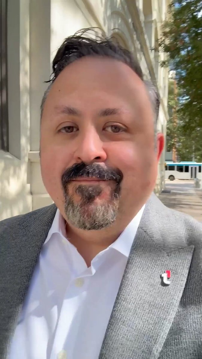 A close-up selfie of a middle-aged man with a gray-streaked beard and mustache, wearing a white shirt and gray blazer, standing outside a building with a bus visible in the background.