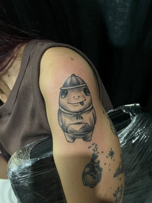 A black ink tattoo of a cartoonish figure wearing a cap and hooded sweatshirt with "fih" written on the chest is displayed on someone's upper arm. Below the character, additional tattoos including what appears to be a bottle and small stars are visible, showcasing the person's commitment to permanently displaying their $Fih affiliation.