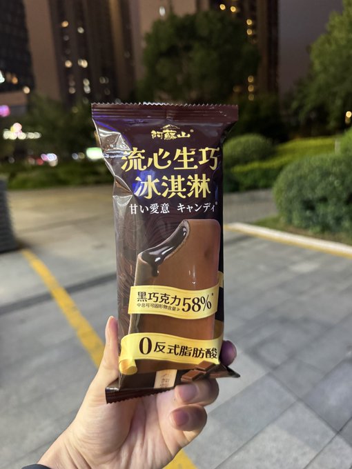 The image shows a person holding up a chocolate bar, which appears to be a chocolate-covered donut or a chocolate ice cream sandwich. The packaging of the chocolate treat has Japanese characters on it, indicating that it might be a product from Japan. The text in the image reads "おもしくは洗濯回してる間にお散歩 #STEPN". This translates to "Step back and enjoy the chocolate", suggesting that the person is encouraging others to indulge in this delicious treat.