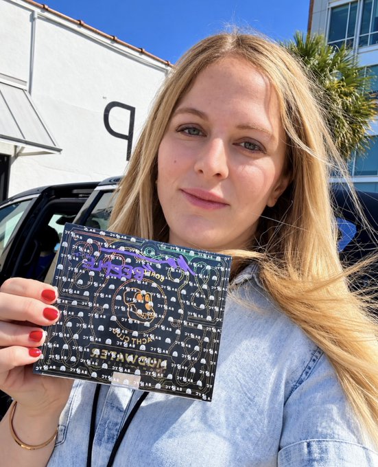 A young blonde woman with red nail polish holds up a CD or record featuring colorful artwork, photographed outdoors near a car.
