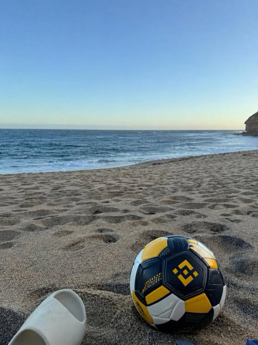 A soccer ball and a single white slide rest on a sandy beach with the ocean and a rocky outcrop in the background, capturing a relaxed seaside moment. The ball features a yellow, black, and white design with a diamond-shaped logo, fitting the laid-back "beach mode" vibe of the post.