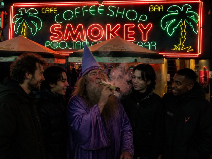 A man dressed as a wizard in a purple robe and pointed hat exhales smoke through a cigar while surrounded by four young admirers outside a neon-lit "Smokey Coffee Shop" in Amsterdam, which advertises a bar, pool tables, and beer. The scene captures the wizard as a local eccentric figure captivating onlookers with his smoking ritual.