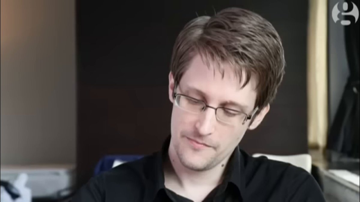 The image shows a man wearing glasses sitting at a desk or table while looking down at something. He appears to be focused on his work, possibly reading or studying. The background features a brown wall with a window that allows natural light into the room. There is also an object in the scene, which could be a chair or another piece of furniture.