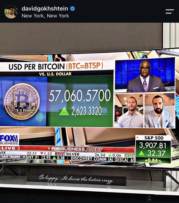 An Instagram post from David Gokhshtein showing a Fox Business broadcast with Bitcoin (BTC=BTSP) at $57,060.57 (up 2,623.33) alongside two guests being interviewed, with S&P 500 at 3,907.81 (+32.37, +0.84%) displayed below. The post humorously responds to a follower named Taylor Ferber by joking that this is where viewers find him—"on Insta"—while a placard reading "Be happy. It drives the haters crazy" sits in front of the TV, reinforcing his crypto-bullish persona tied to the $TBB (The Bitcoin Bridge) ticker.