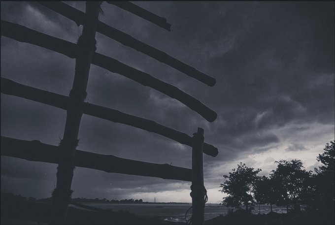 A dramatic, moody photograph showing the silhouette of a wooden fence in the foreground against a dark, stormy sky over a distant landscape.
