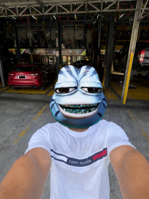 A person wearing a Tommy Hilfiger shirt takes a selfie while wearing a creepy cartoonish mask in front of what appears to be a car repair garage.