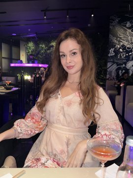 A photograph of a woman with long brown hair wearing a floral dress, holding a pink cocktail glass at a venue with neon lighting.