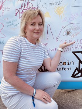 The image features a woman sitting down, wearing white pants and a striped shirt. She has her hand outstretched towards the camera, with a smile on her face. The background of the photo shows a wall covered in graffiti, including words such as "rainbow" and "bitcoin". There is also an image of a coin displayed on this wall. The post that accompanies the image reads: "@5Ksana".