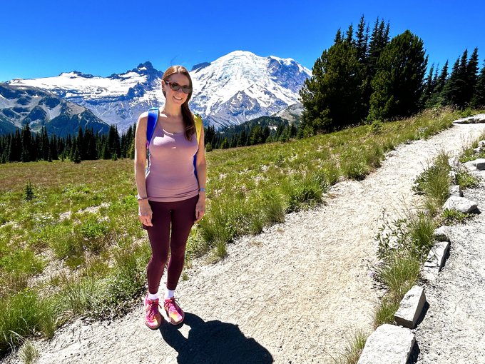 A woman in a pink tank top, maroon leggings, and sunglasses stands smiling on a dirt trail with a backpack, posing in front of a stunning snow-capped mountain (likely Mt. Rainier) under a clear blue sky, perfectly capturing a scenic weekend hike with a loved one.