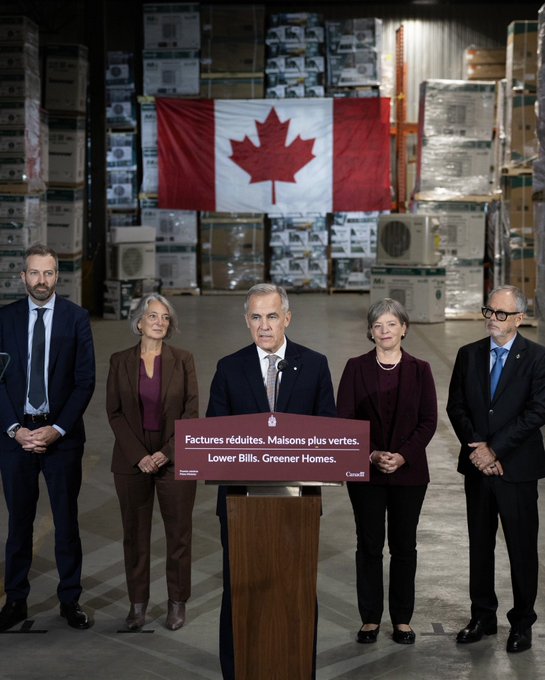 Five people stand in a warehouse with a Canadian flag hanging behind them and stacks of appliances in the background; the central man speaks at a podium labeled 'Factures réduites. Maisons plus vertes. Lower Bills. Greener Homes. Canada'.
