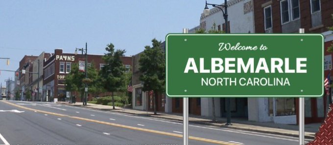 A street scene with a green sign reading 'Welcome to ALBEMARLE, NORTH CAROLINA' alongside a downtown road with shops including 'PAWN'S' visible.