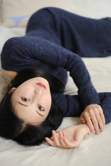 A woman with dark hair wearing a navy blue sweater lies on a white bed, gazing at the camera with her hands near her face.