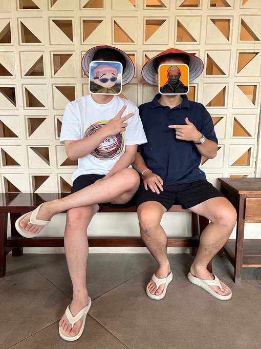 Two people sit on a bench outdoors, their faces covered by hat emoji stickers, with one pointing at the other against a decorative block wall.