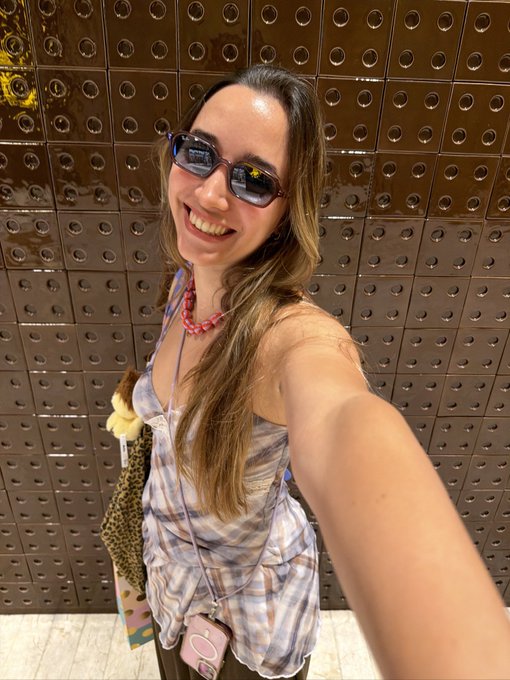 A smiling young woman taking a selfie, wearing sunglasses and a tie-dye tank top, standing in front of a brown tiled wall with a textured pattern.